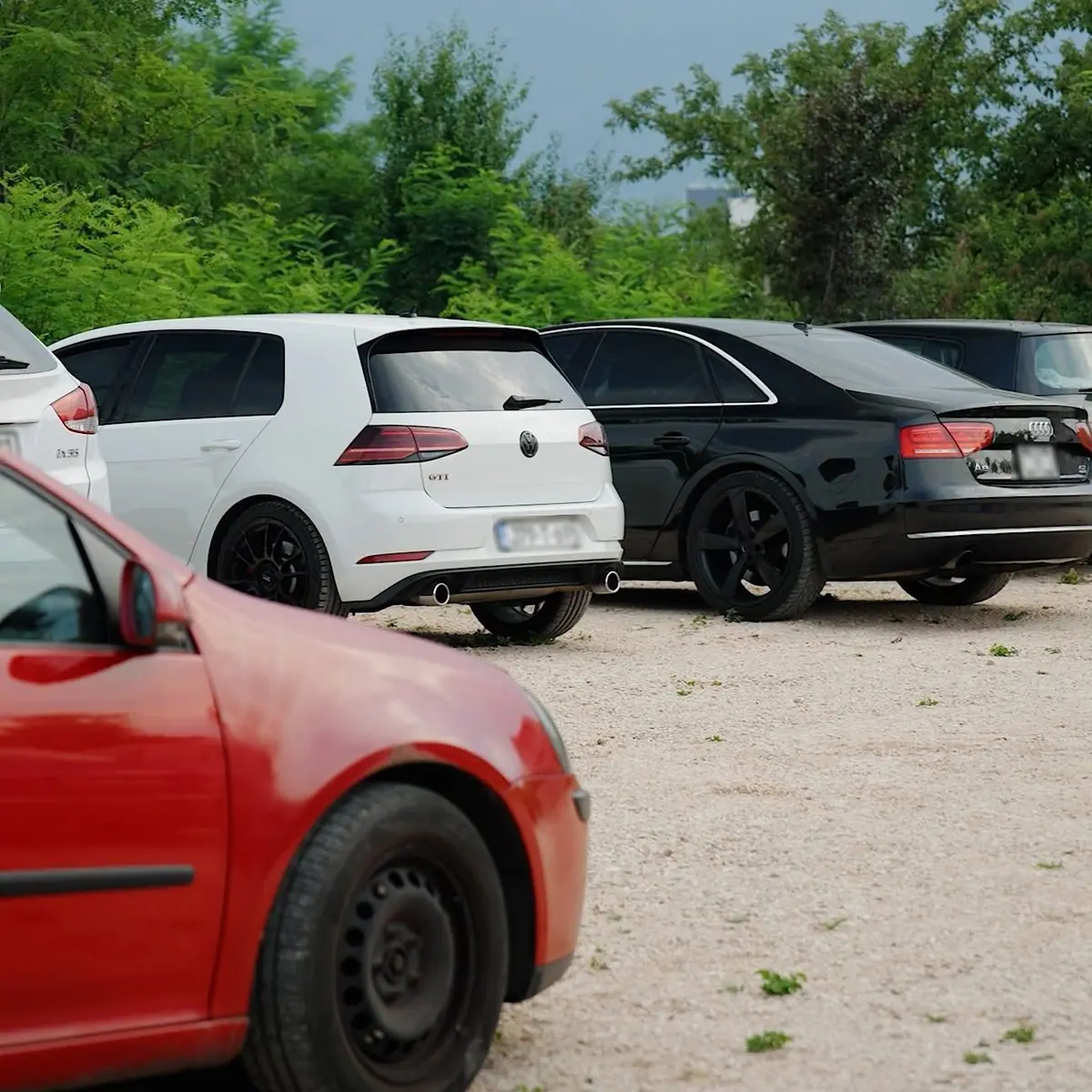 Bijeli Golf i crni Audi parkirani na parkingu, okruženi zelenilom