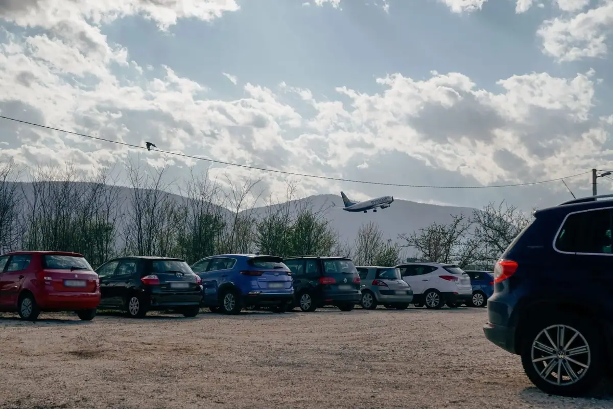Avion polijeće iznad automobila parkiranih na parkingu Sky Parking & Fly, a iza njih se vide planine